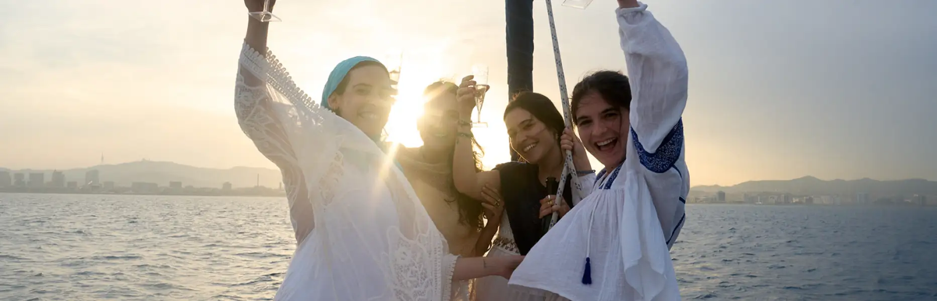 Freundinnengruppe stößt bei Sonnenuntergang auf einem Boot vor der Skyline von Barcelona mit Cava an