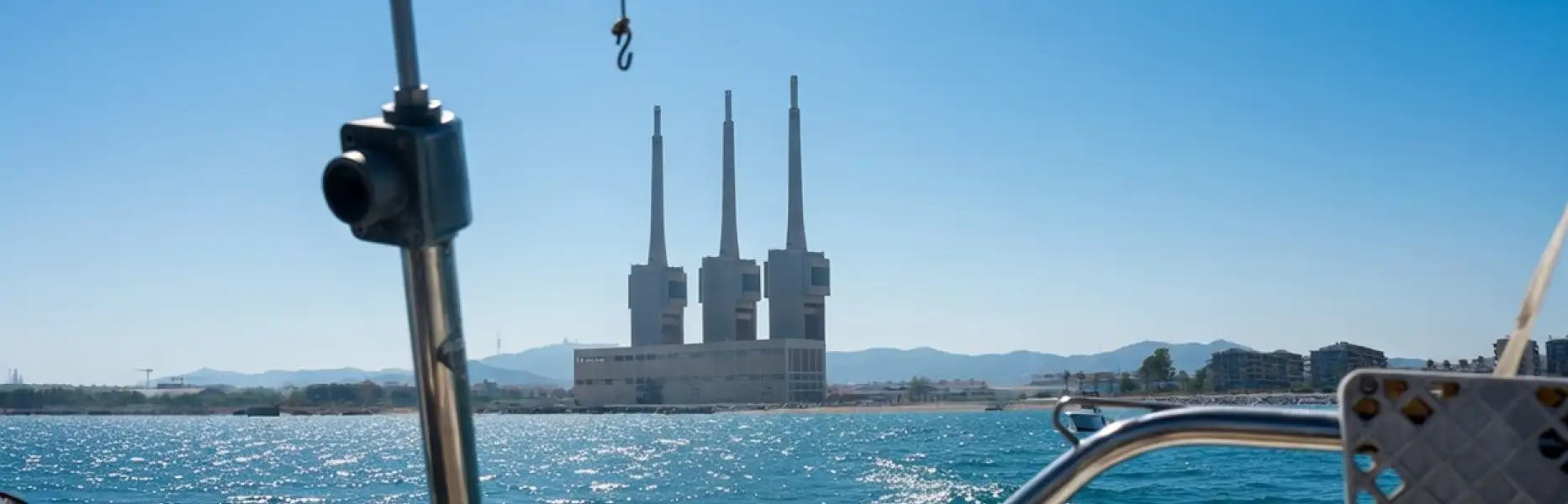 Vista desde un velero navegando frente a la costa de Barcelona con el mar azul y las torres de la central de Sant Adrià al fo