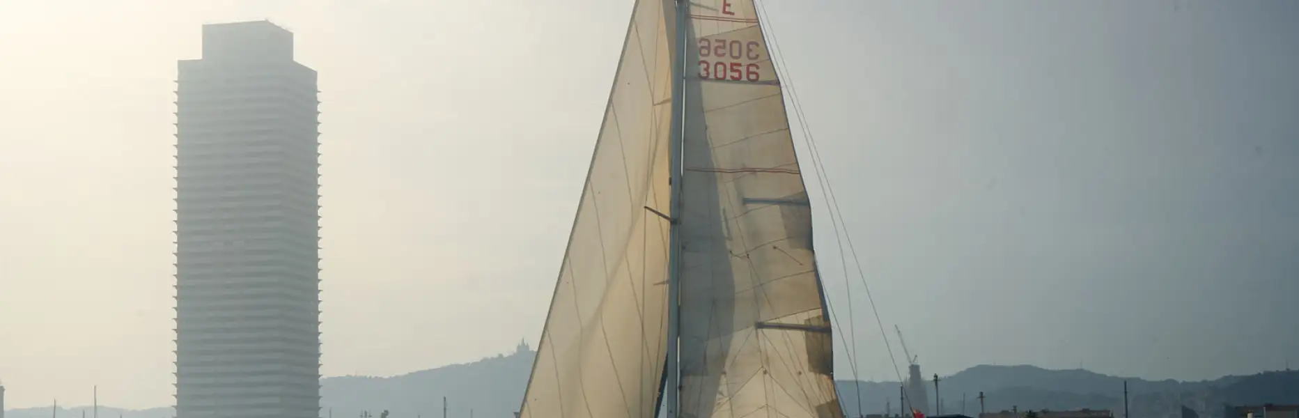 Velero de charter navegando al atardecer frente al puerto y los edificios de Barcelona