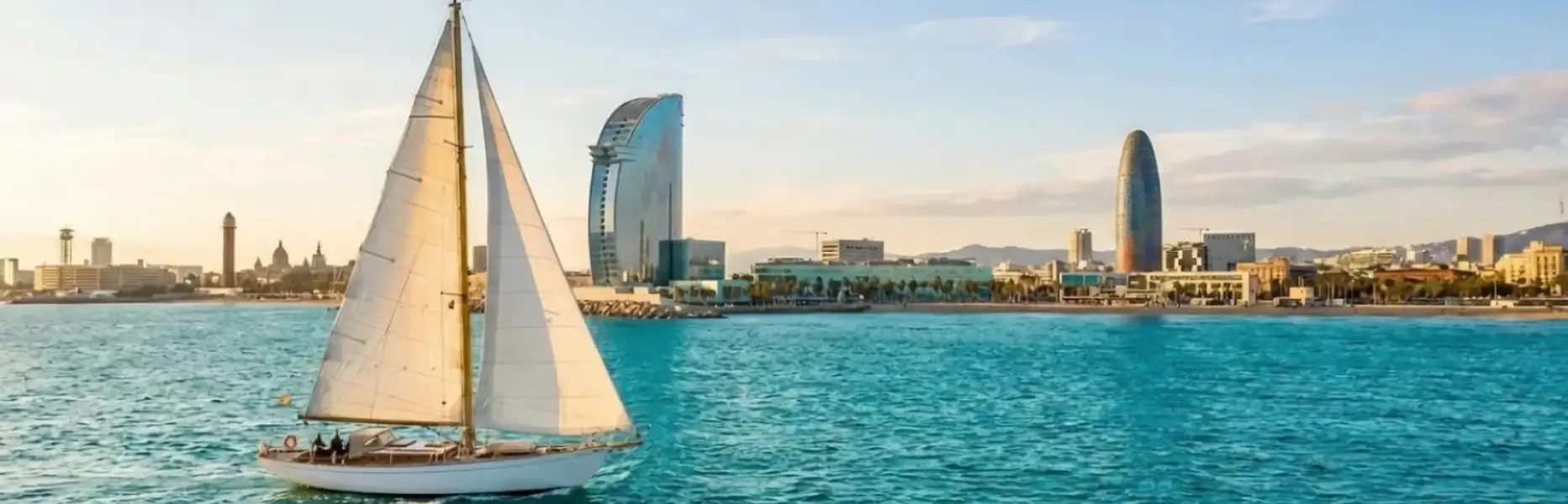 Barca a vela a noleggio che naviga al tramonto davanti allo skyline marittimo di Barcellona