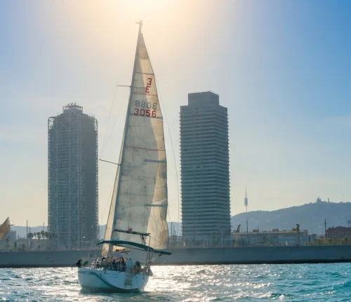 Velero navegando frente al skyline de Barcelona con sol bajo y mar en calma